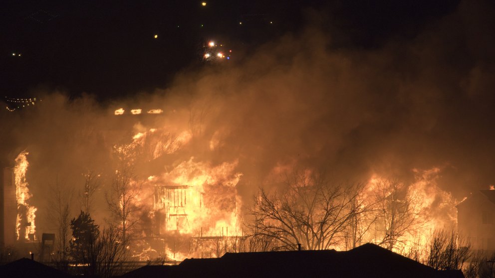A wildfire at night burns orange, engulfing buildings and surrounding vegetation.