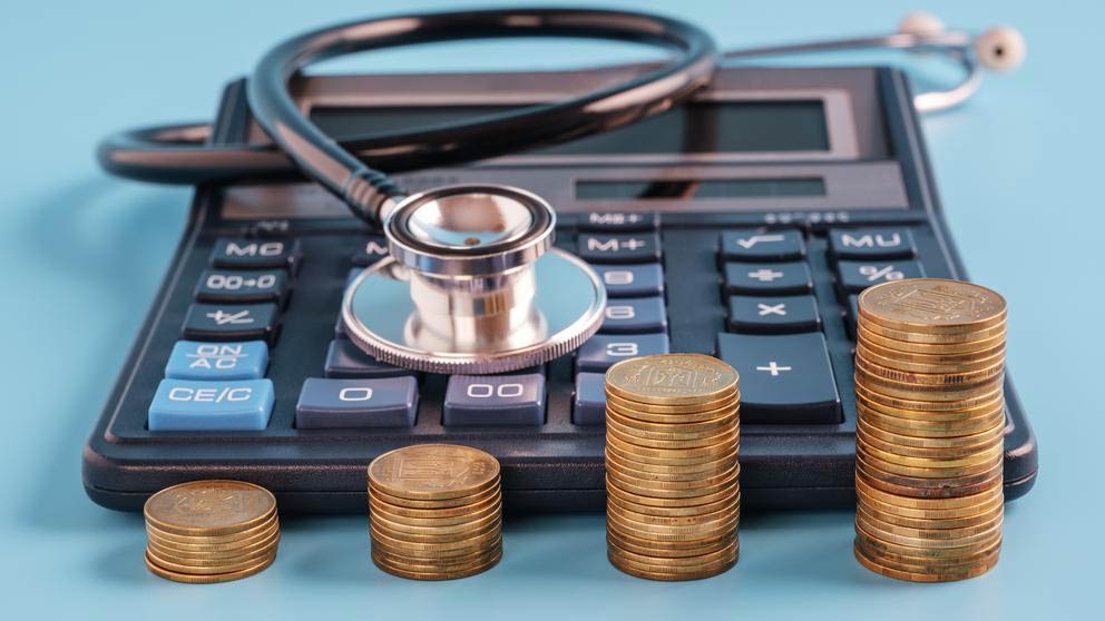 A black calculator with a stethoscope coiled on top and 4 stacks of coins in front of it. The coin stacks increase in height from left to right.